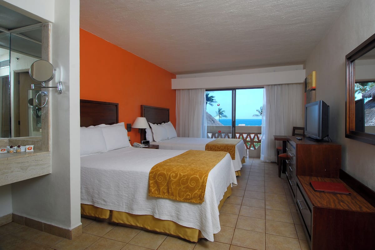 A hotel room with two beds, a TV, a desk, and a balcony with a view of the ocean.