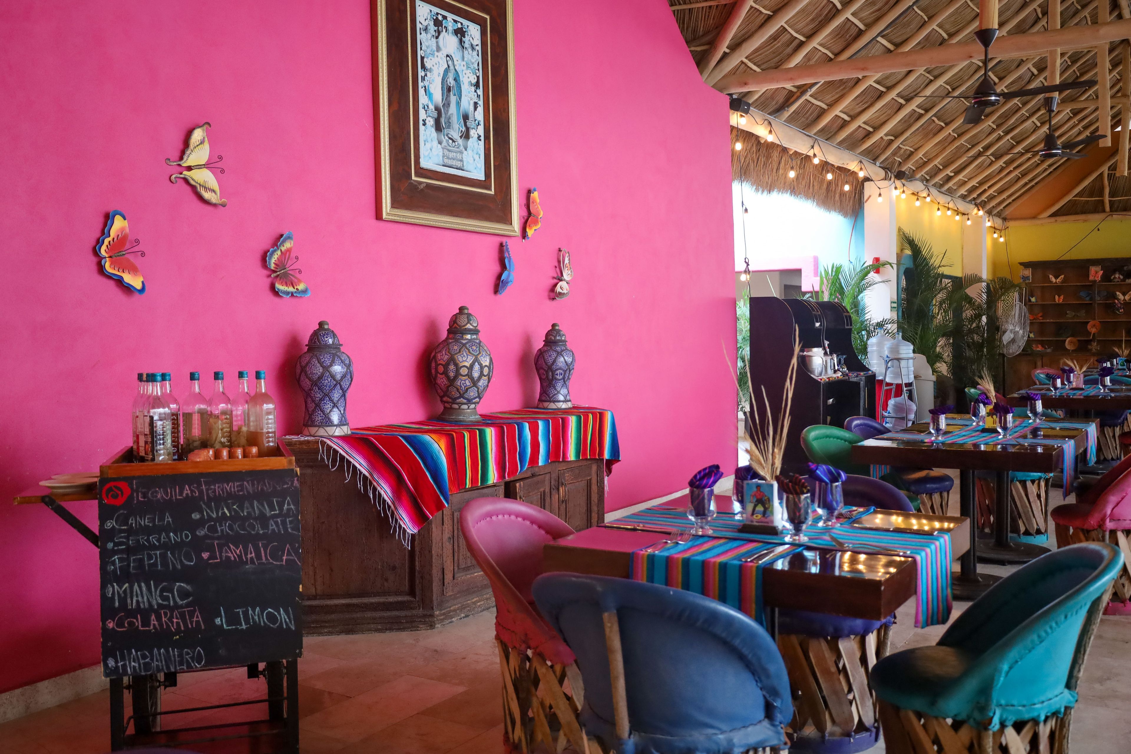 Interior de un restaurante con una pared rosa vibrante, mobiliario de madera y techo de paja.