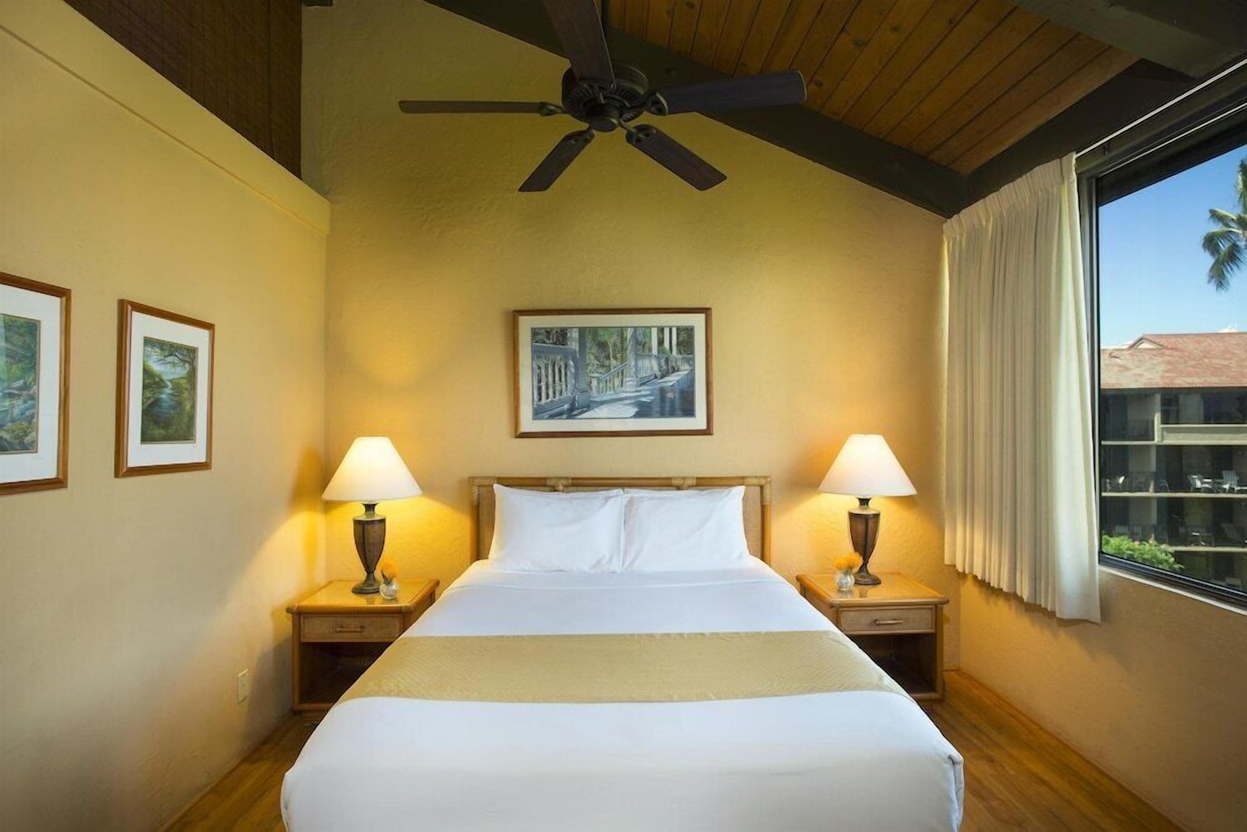 Une chambre d’hôtel avec un lit, deux tables de chevet avec des lampes, un ventilateur de plafond, un tableau encadré au mur et une fenêtre avec des rideaux.