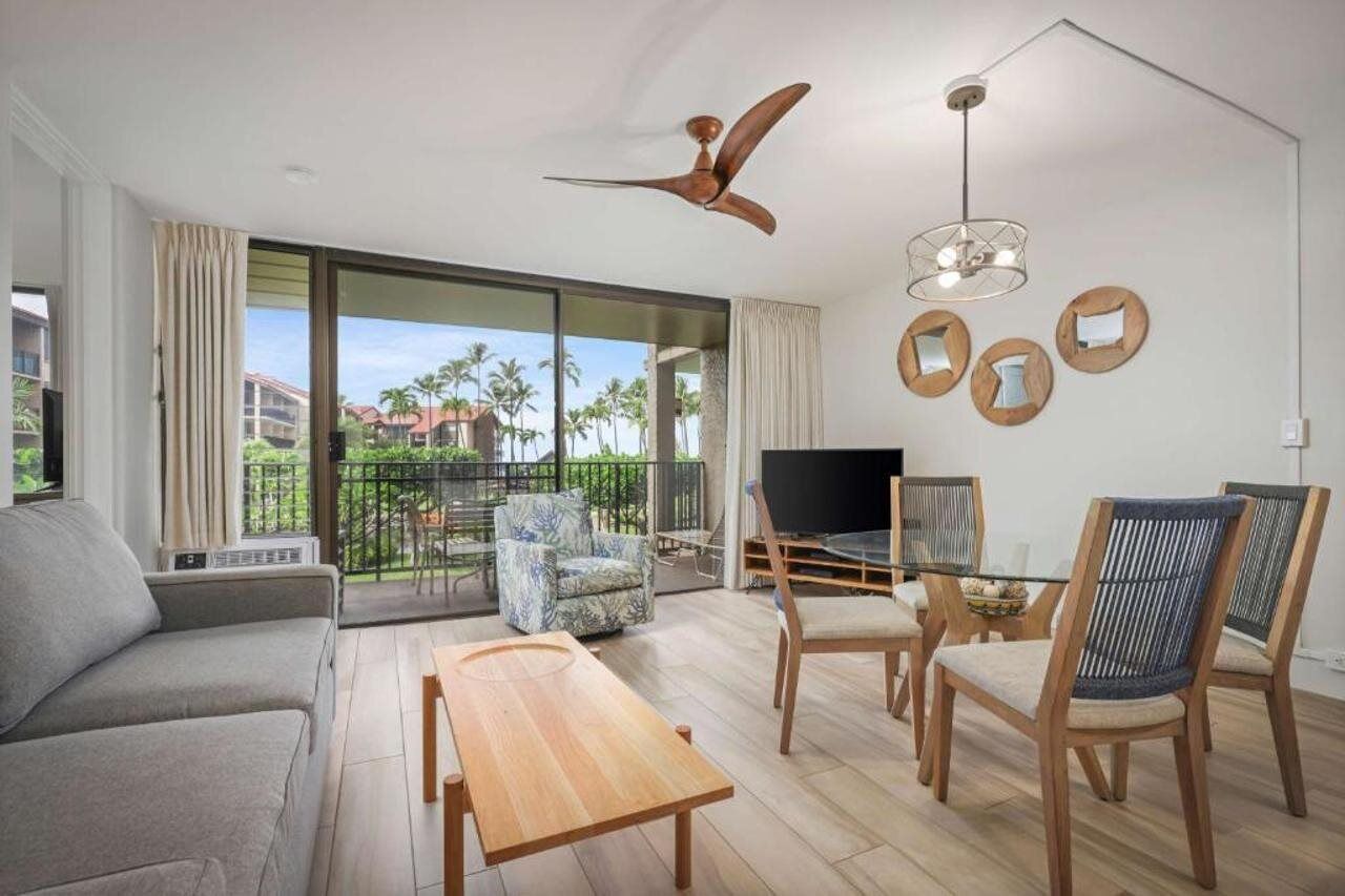 A modern living room with a sofa, a wooden coffee table, a TV, and a balcony with a view of palm trees.