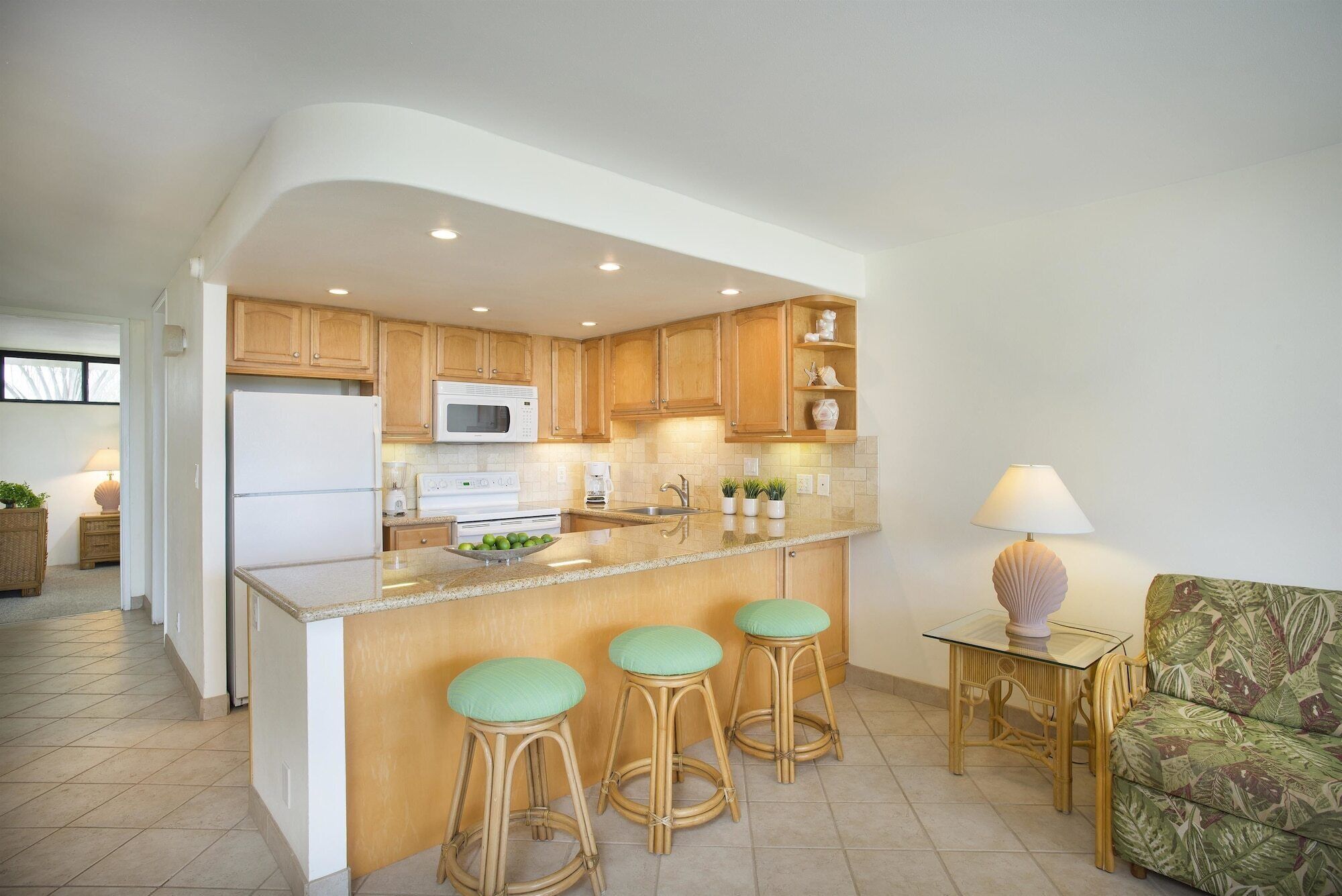 A modern kitchen with wooden cabinets, a white refrigerator, a microwave, and a sink. There are three bar stools with green cushions and a small table with a lamp.