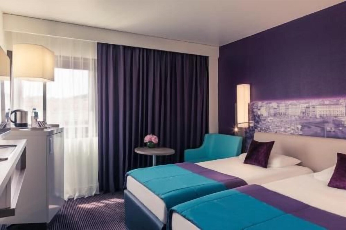 A hotel room with a bed, a desk, a chair, a small table, and a wall with a cityscape mural.