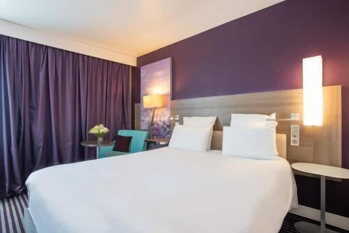 Une chambre d’hôtel avec un grand lit, une petite table avec une lampe, une chaise et une vue sur le paysage urbain.