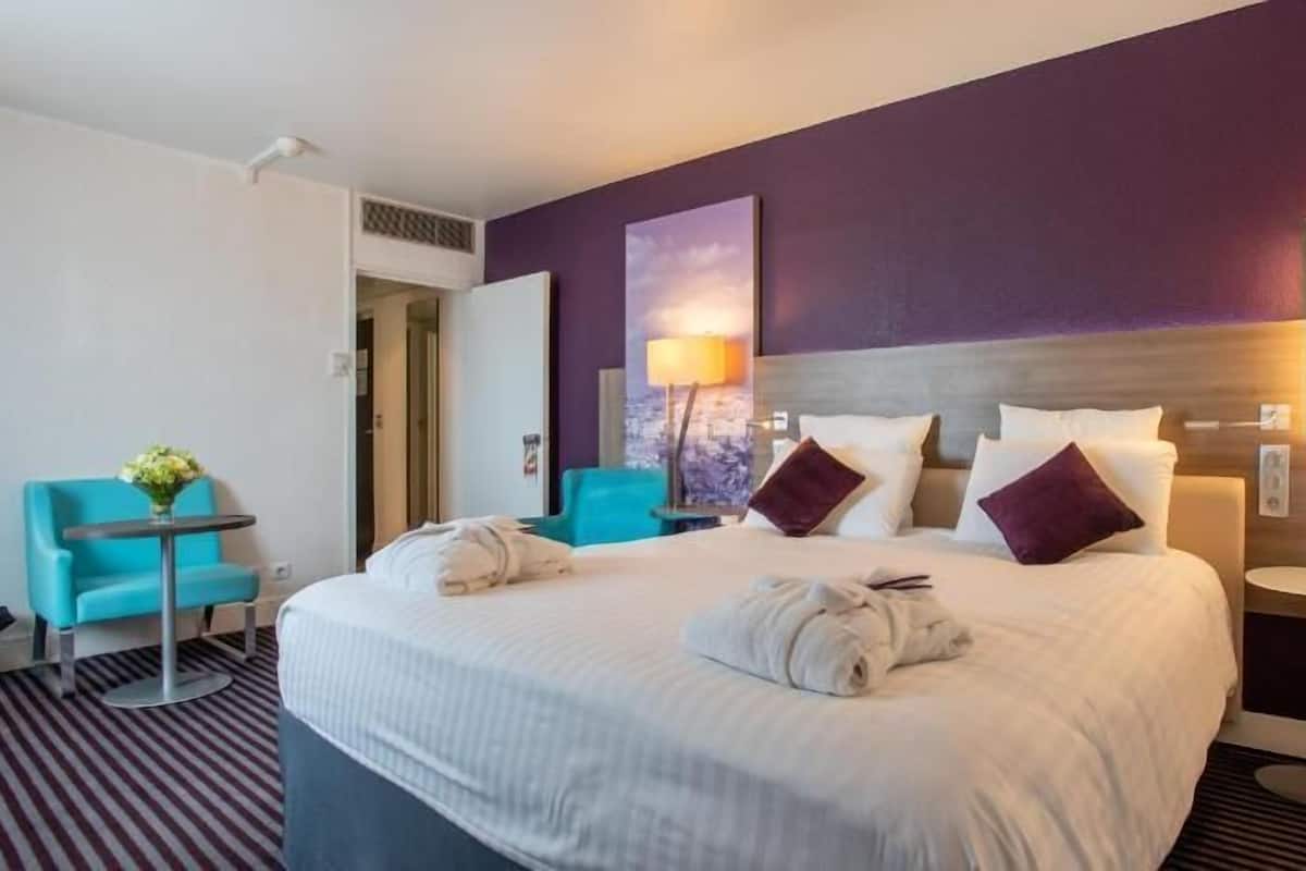 Une chambre d’hôtel avec un grand lit, une petite table, une chaise et une lampe fixée au mur.