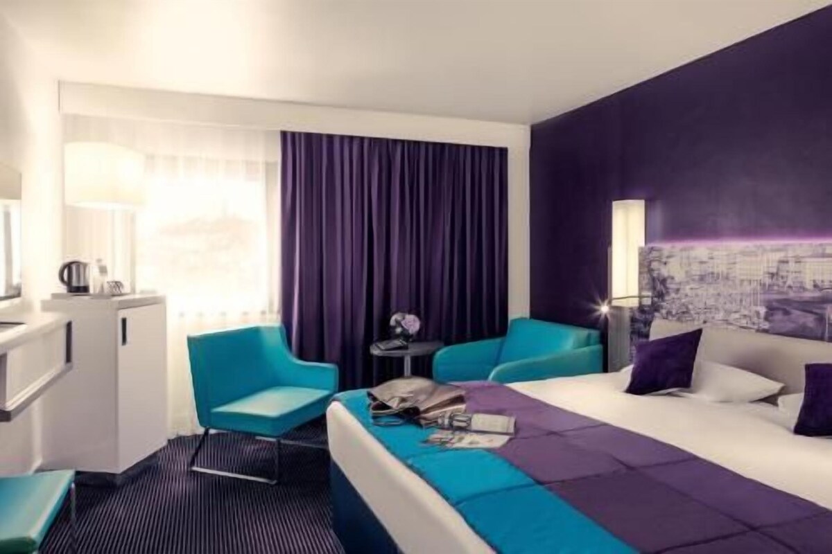 A hotel room with a bed, two chairs, a small table, a lamp, and a cityscape painting on the wall.