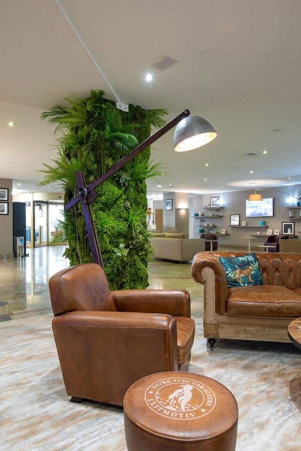 A lobby with a leather armchair, a sofa, and a round ottoman with a logo. A tall plant and a lamp are also visible. - Hôtel Mercure Marseille Centre Vieux-Port (MARSEILLE)