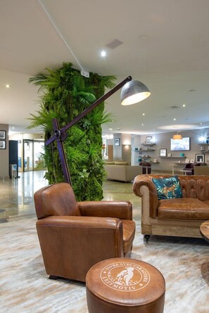 A lobby with a leather armchair, a sofa, and a round ottoman with a logo. A tall plant and a lamp are also visible. - Hôtel Mercure Marseille Centre Vieux-Port (MARSEILLE)