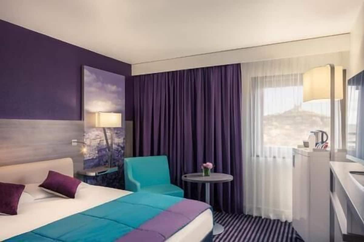 Une chambre d’hôtel avec un lit, un bureau, une chaise, une petite table avec un vase et une fenêtre avec des rideaux.