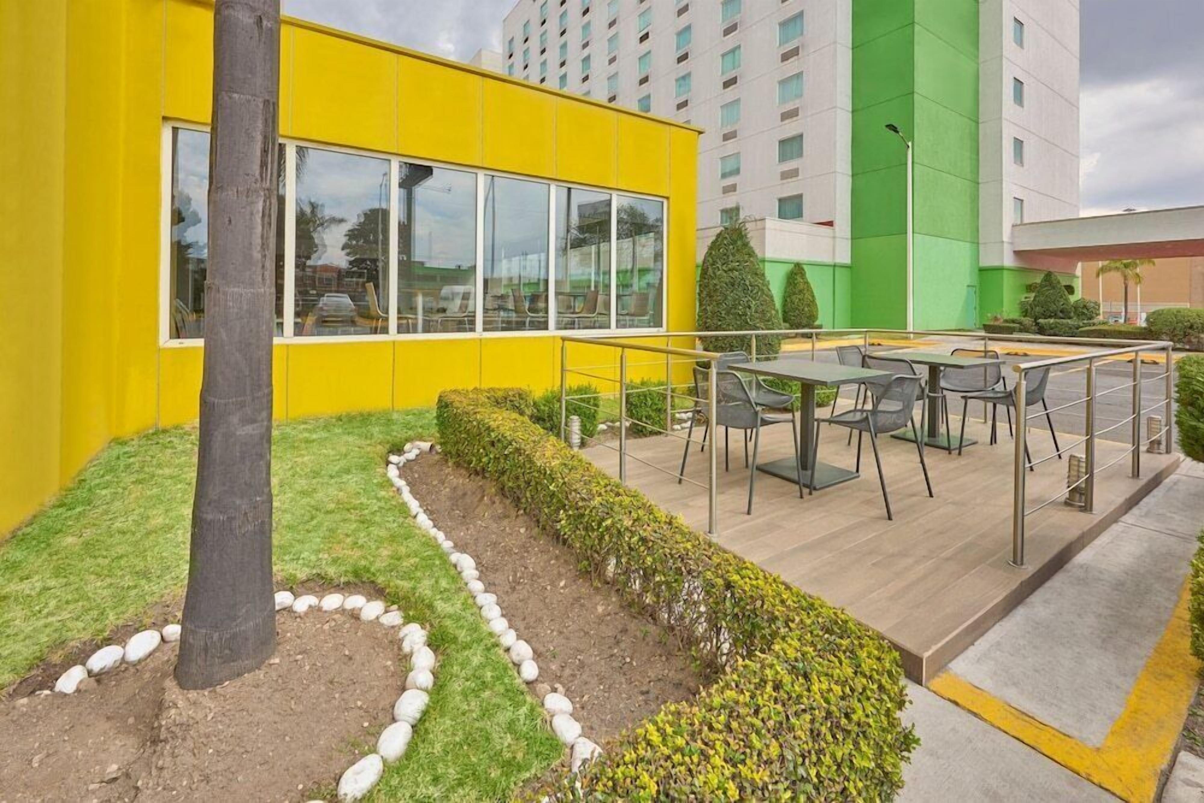 A modern building with a yellow facade, large windows, and outdoor seating area with tables and chairs.