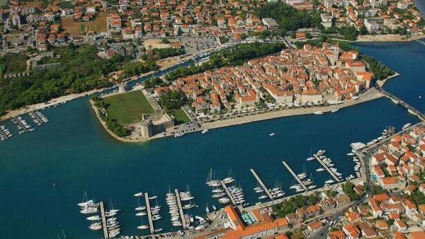 Vista aérea de uma cidade costeira com um porto, áreas residenciais e uma estrutura imponente que lembra um castelo.