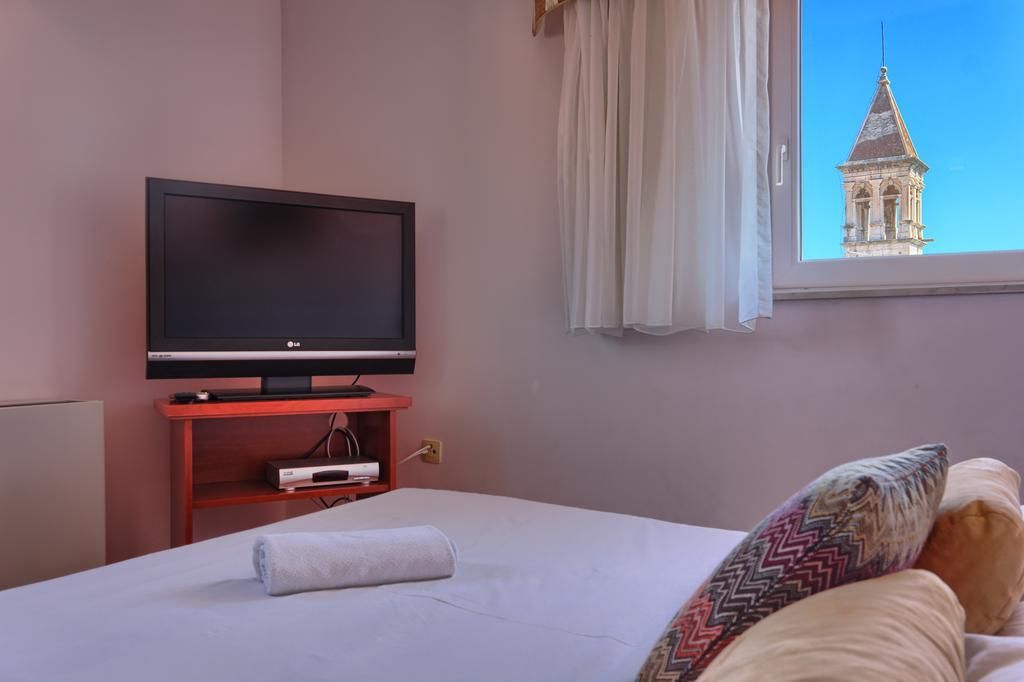 A hotel room with a bed, a television on a stand, a window with a view of a building, and a curtain.