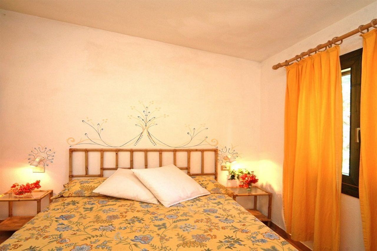 Una camera da letto con una coperta a fiori, una testiera di legno con motivo a forma di albero, due comodini con lampade e una finestra con tende arancioni.