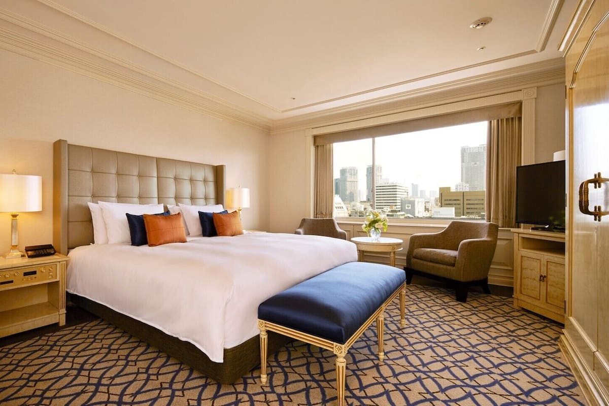 A hotel room with a large bed, a desk, a chair, a TV, and a window with a city view.