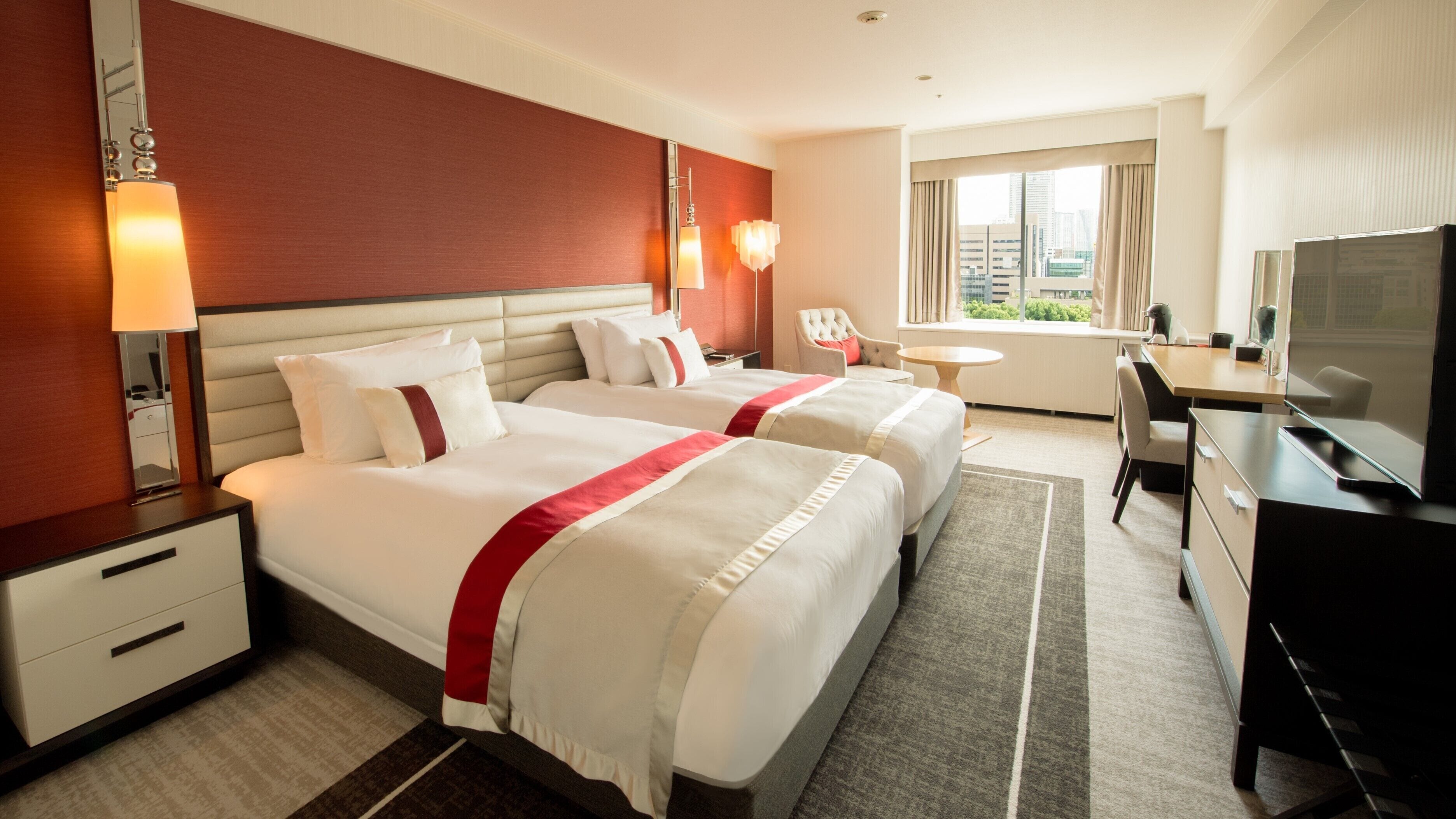 A hotel room with two beds, a desk, a chair, a TV, and a window with a city view.