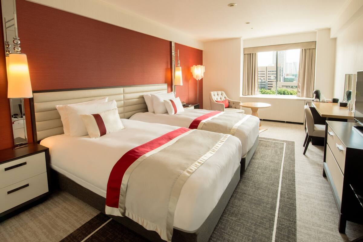 A hotel room with two beds, a desk, a chair, a TV, and a window with a city view.