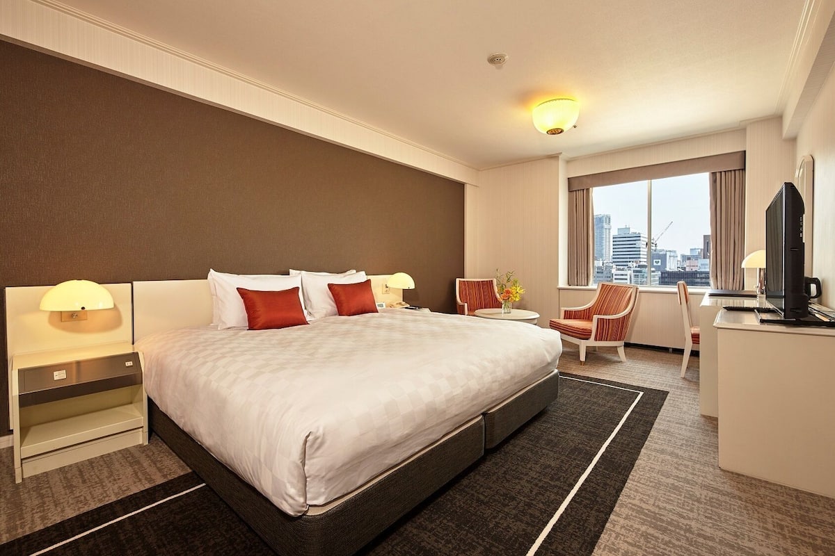 A hotel room with a large bed, a desk with a computer, a chair, and a view of the city.