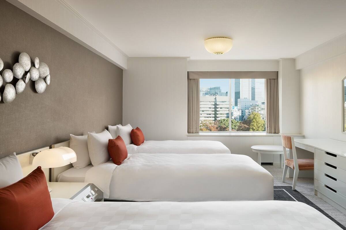 A hotel room with two beds, a desk, a chair, a TV, and a large window with a city view.
