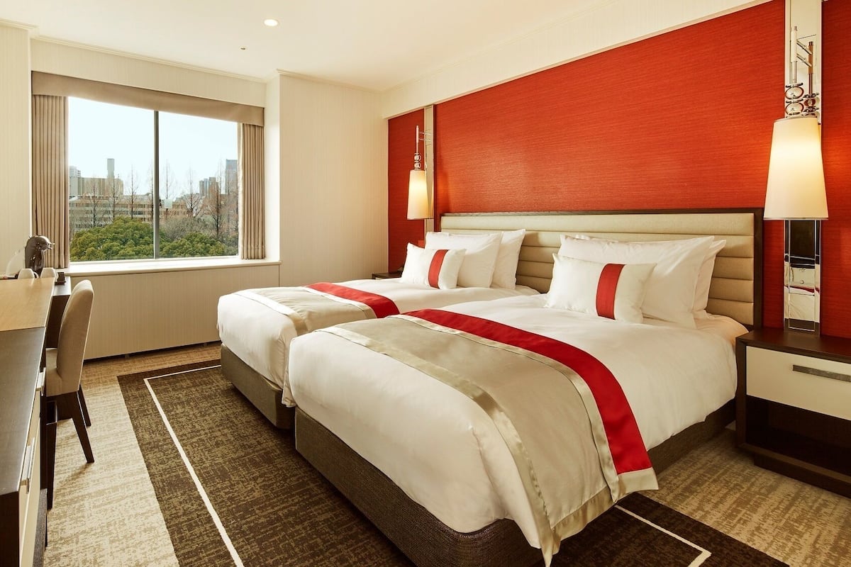 A hotel room with two beds, a desk, a chair, a window with a city view, and a red accent wall.