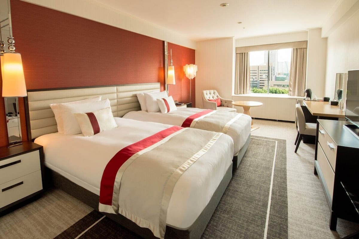 A hotel room with two beds, a desk, a chair, a TV, and a window with a city view.
