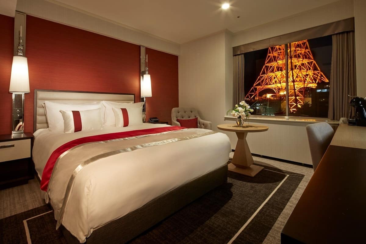 A hotel room with a large bed, a round side table, a chair, a desk, a lamp, and a view of a lit structure outside the window.