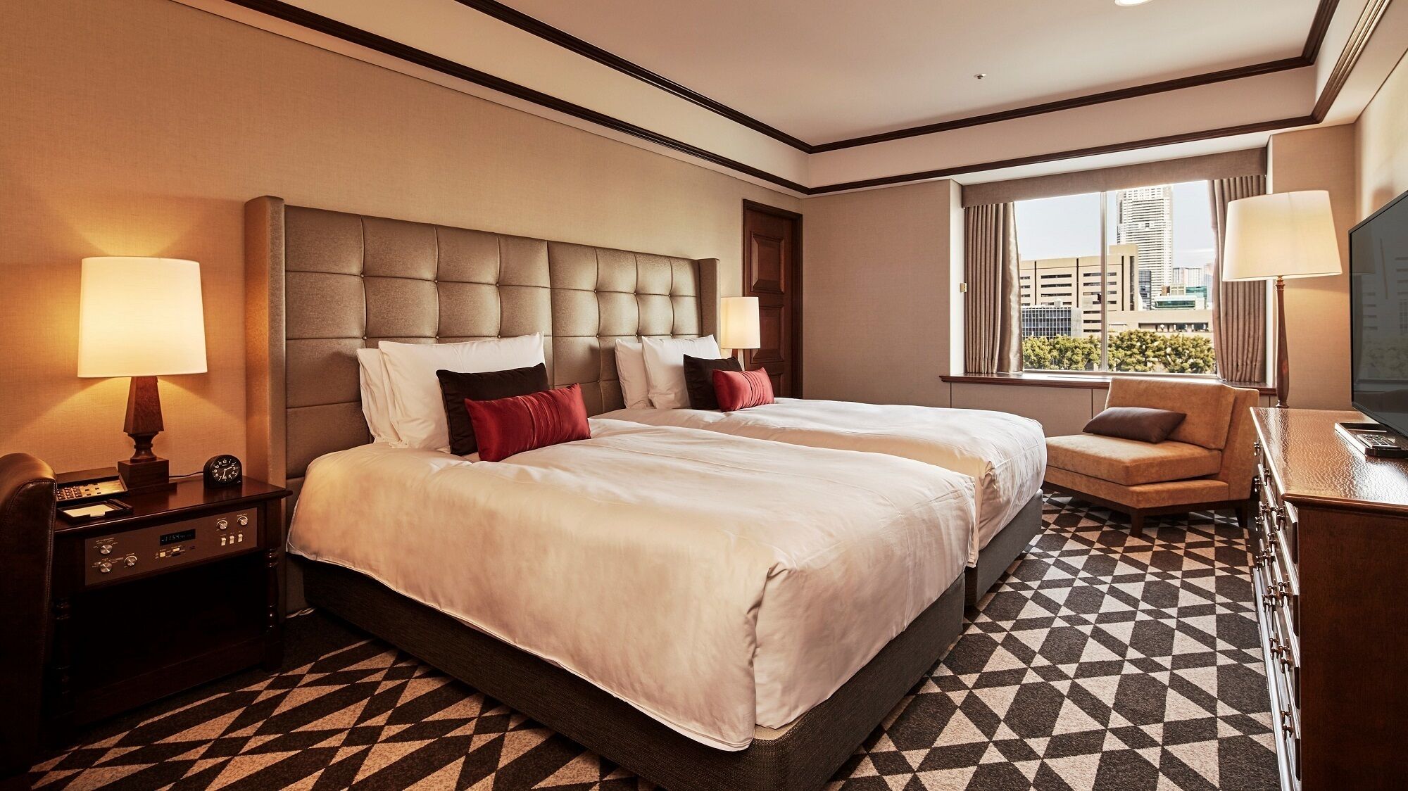 A hotel room with a large bed, two bedside tables, a desk with a chair, a window with a view of buildings, and a patterned floor.