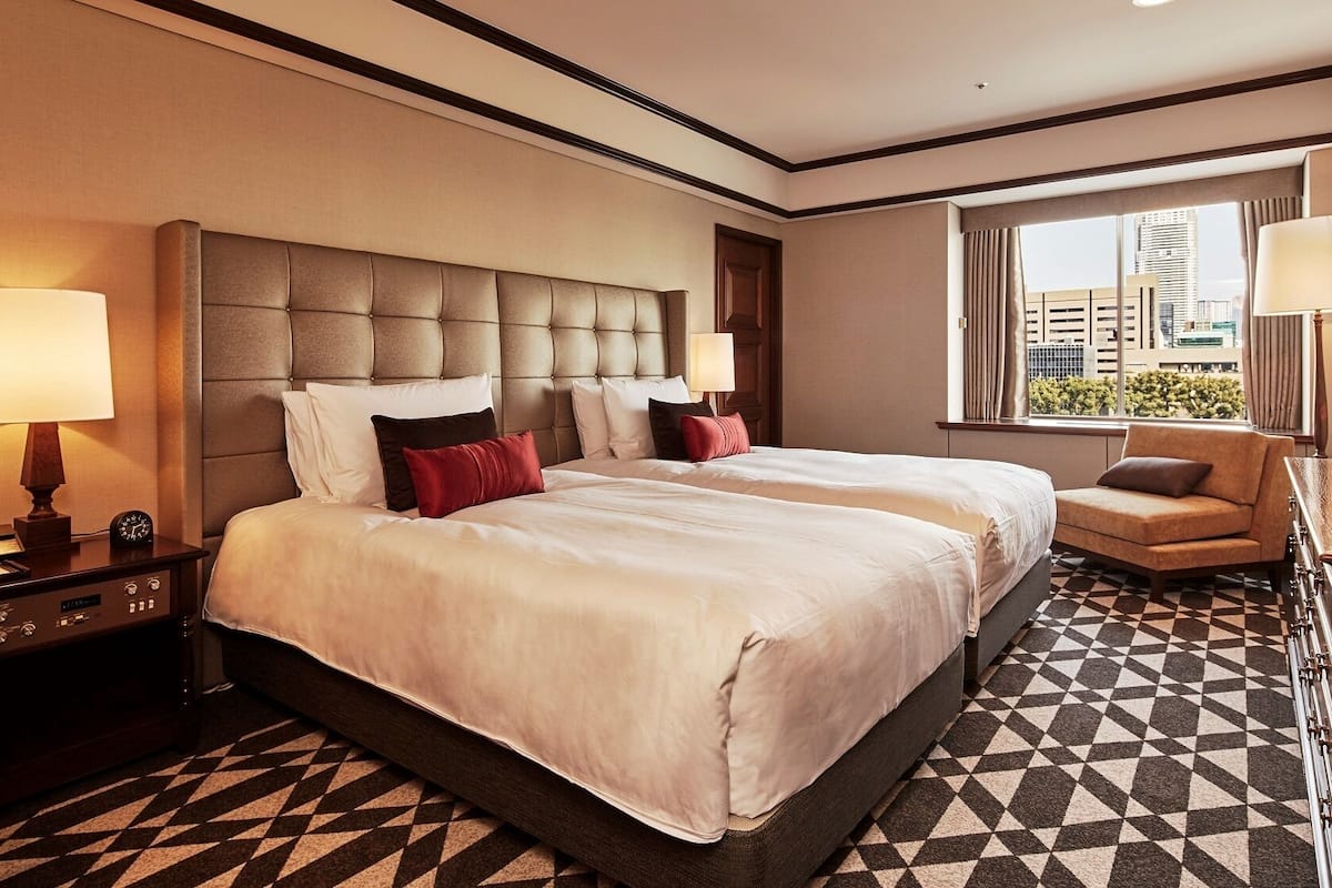 A hotel room with a large bed, two bedside tables, a desk with a chair, a window with a view of buildings, and a patterned floor.