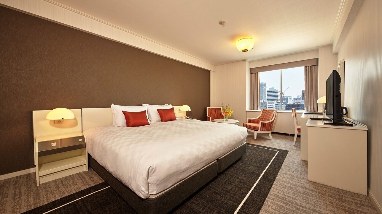 A hotel room with a large bed, a desk, a chair, a TV, and a city view.