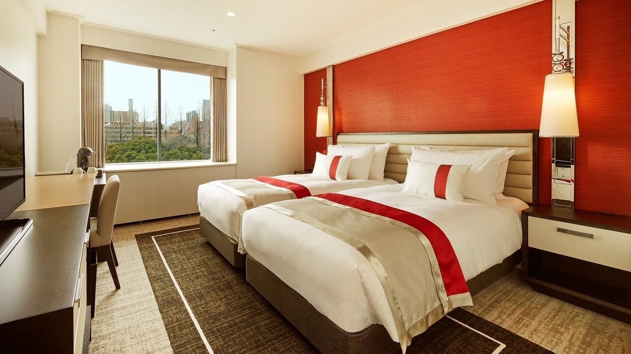 A hotel room with two beds, a desk, a chair, a TV, and a window with a city view.