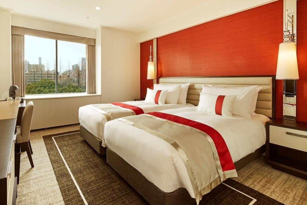A hotel room with two beds, a desk, a chair, a TV, and a window with a city view.