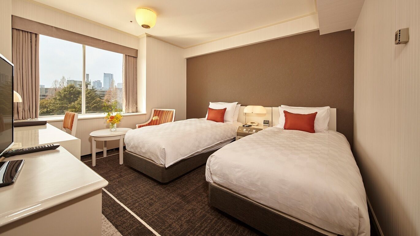 A hotel room with two beds, a desk with a computer, a small table with a vase of flowers, and a large window with a city view.