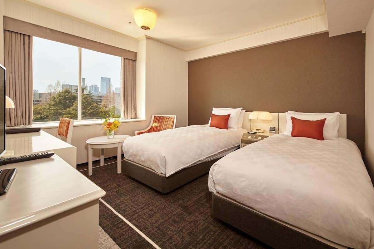 A hotel room with two beds, a desk with a computer, a small table with a vase of flowers, and a large window with a city view.