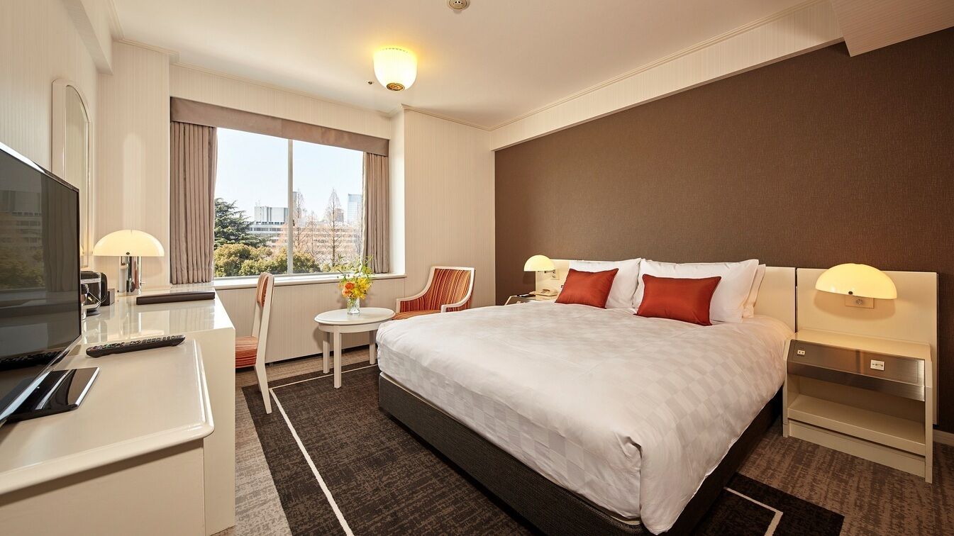 A hotel room with a large bed, a desk with a TV, a small table with a lamp, and a view of the city.