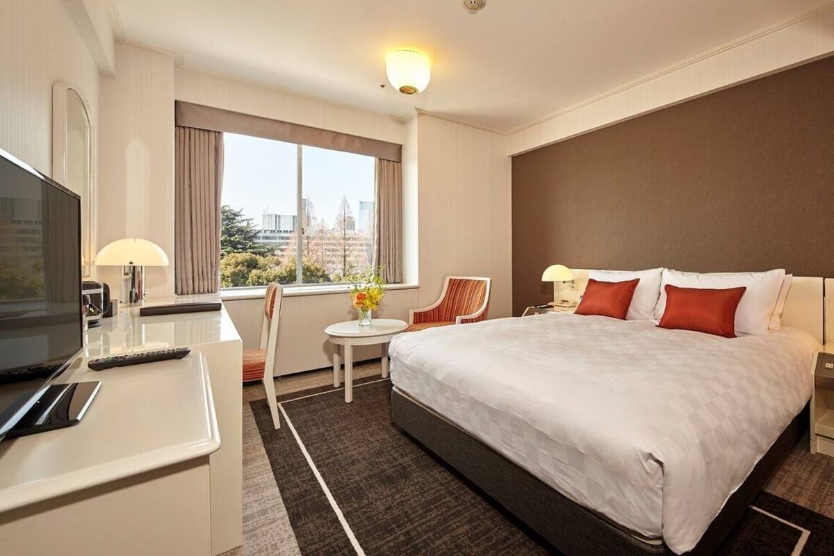 A hotel room with a large bed, a desk with a TV, a small table with a lamp, and a view of the city.