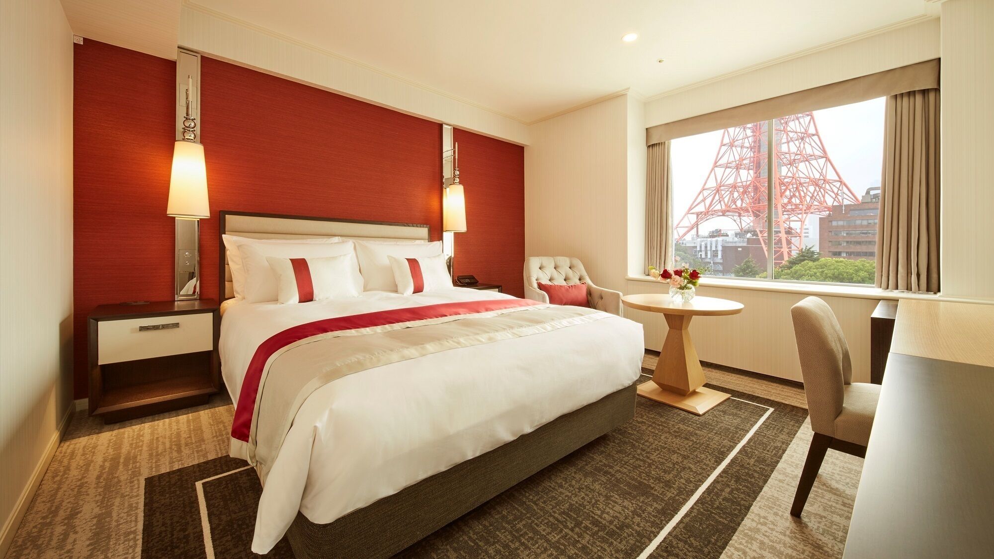 A hotel room with a large bed, a bedside table, a chair, a small round table with flowers, and a window with a view of a red tower.