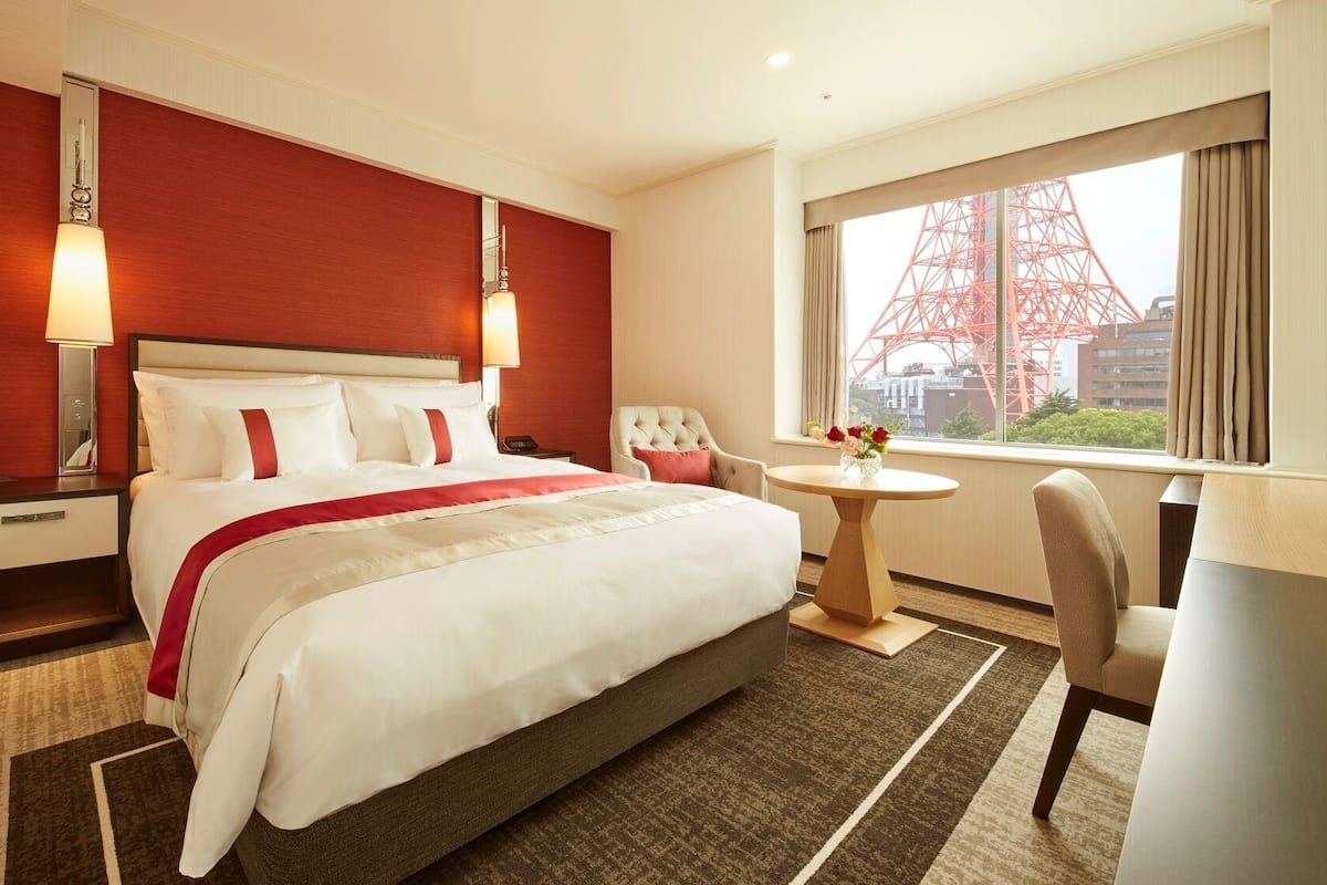 A hotel room with a large bed, a bedside table, a chair, a small round table with flowers, and a window with a view of a red tower.