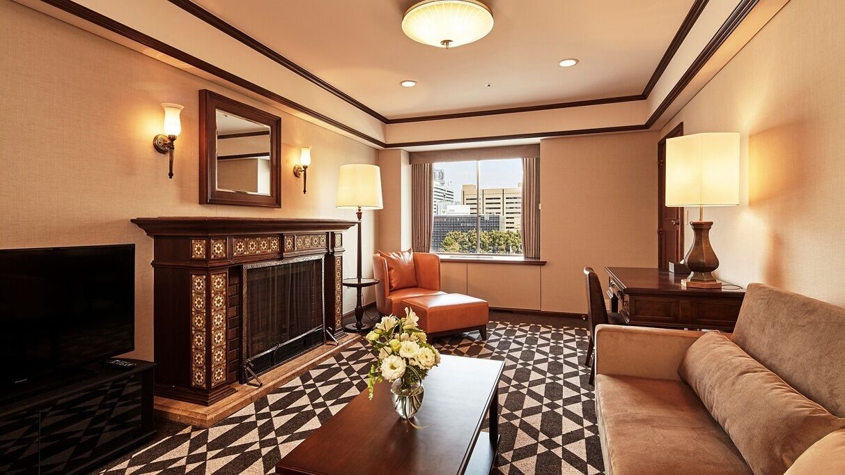 A hotel room with a fireplace, a television, a sofa, a coffee table with flowers, and a window with a city view.