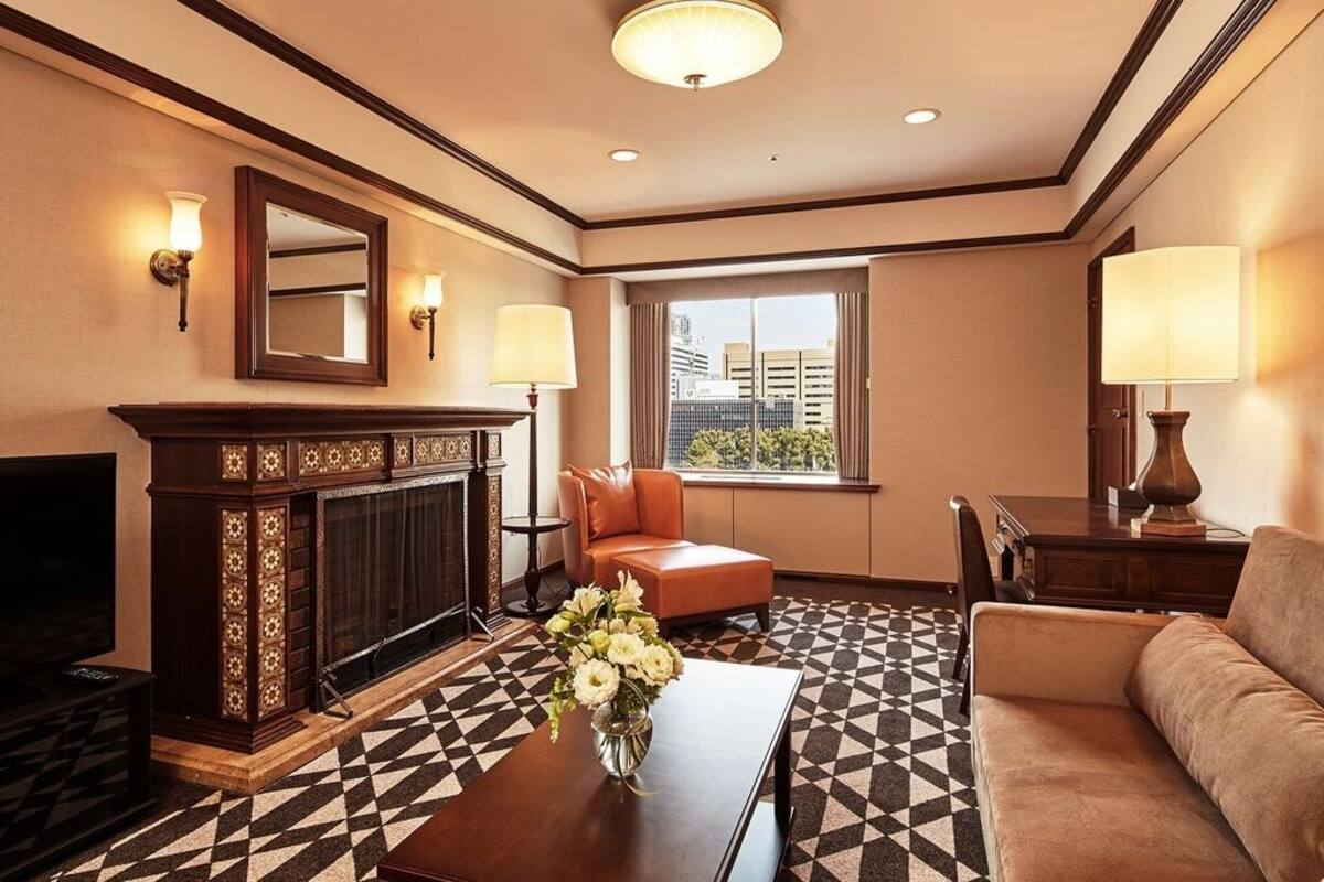 A hotel room with a fireplace, a television, a sofa, a coffee table with flowers, and a window with a city view.