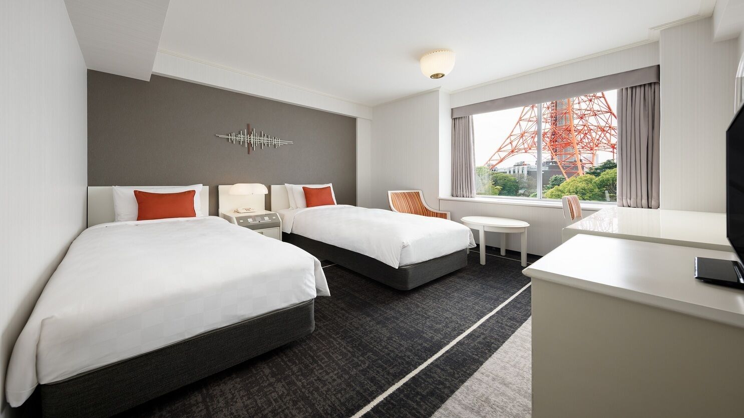 A hotel room with two beds, a desk, a chair, a small table, and a large window with a view of a red structure.