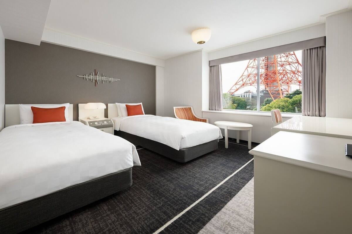 A hotel room with two beds, a desk, a chair, a small table, and a large window with a view of a red structure.