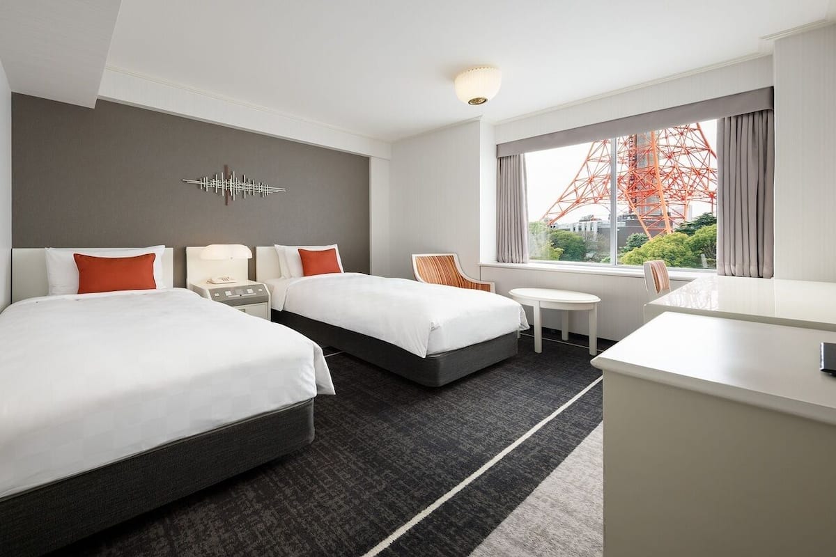 A hotel room with two beds, a desk, a chair, a small table, and a large window with a view of a red structure.