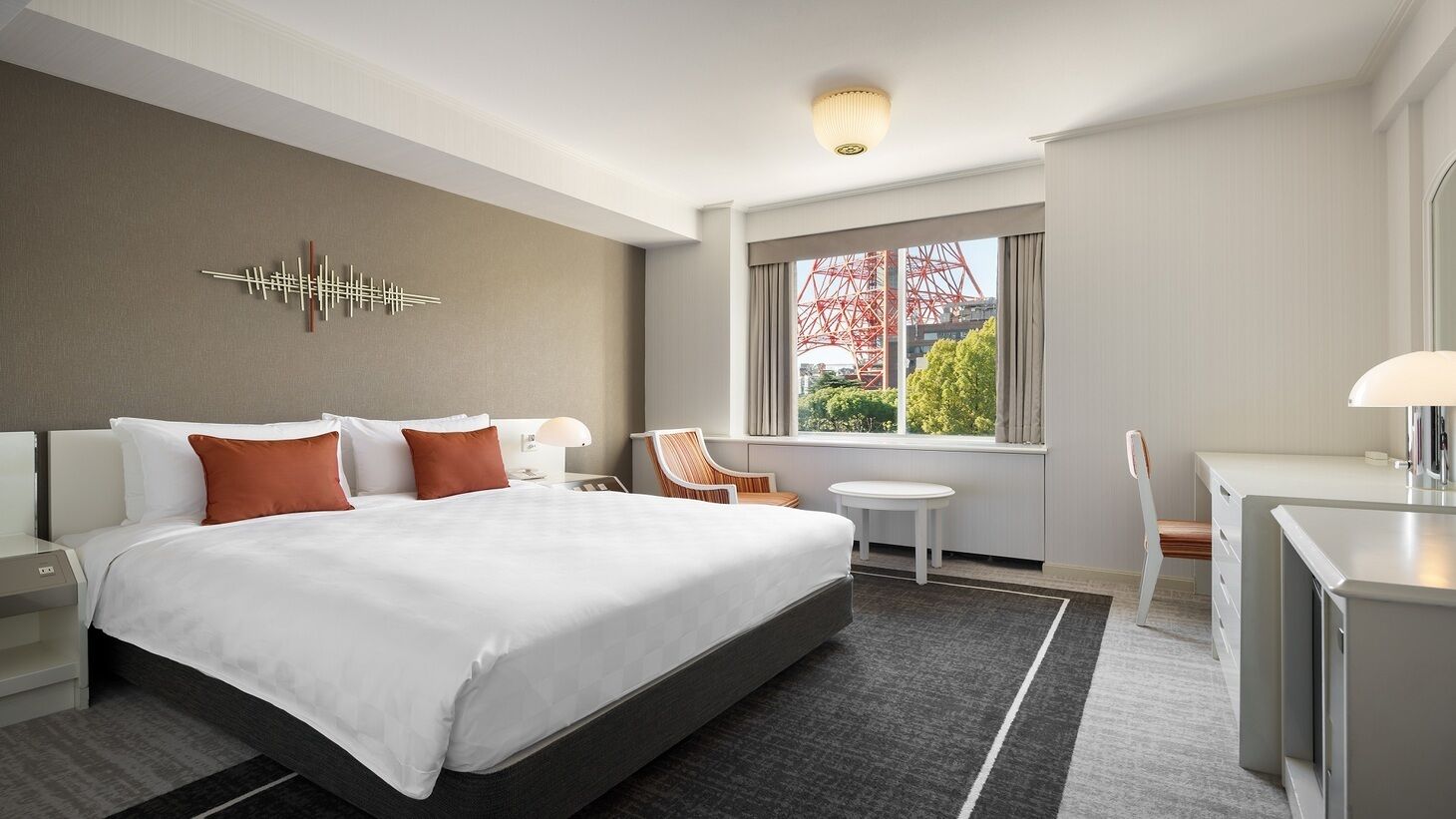 A hotel room with a large bed, a desk, a chair, a small table, and a window with a view of a red structure.