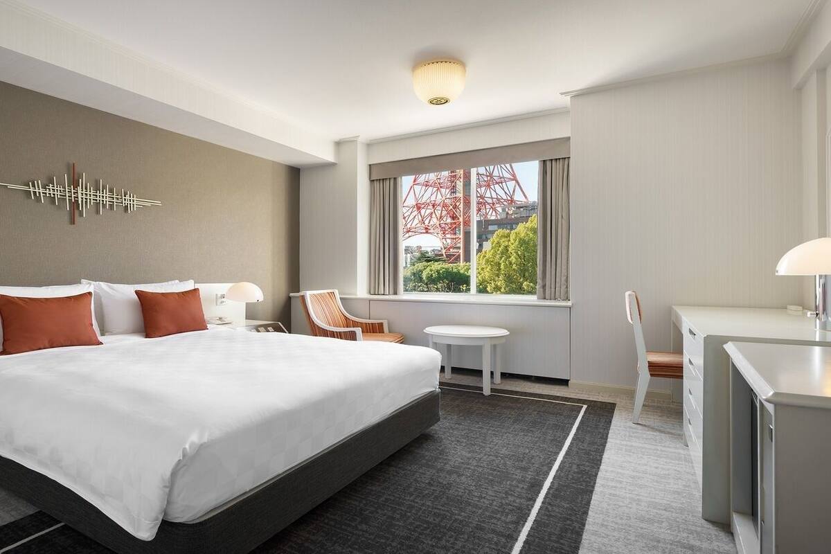 A hotel room with a large bed, a desk, a chair, a small table, and a window with a view of a red structure.