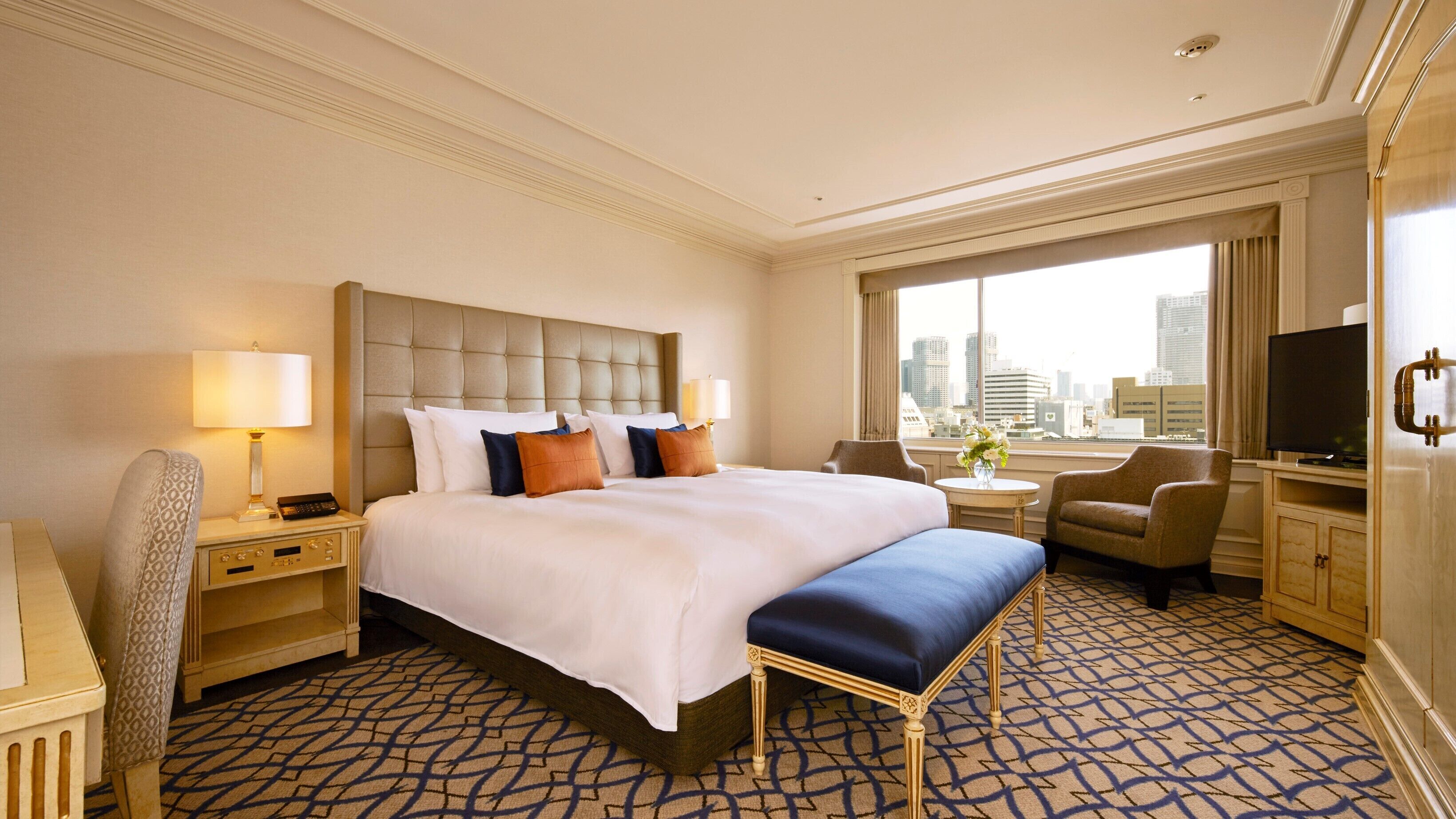 A hotel room with a large bed, a desk, a chair, a TV, and a view of the city.