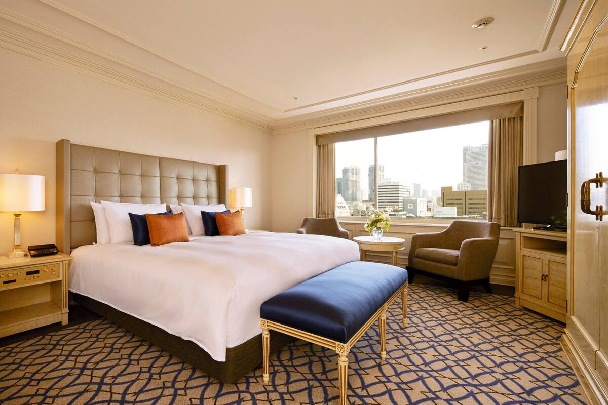 A hotel room with a large bed, a desk, a chair, a TV, and a view of the city.