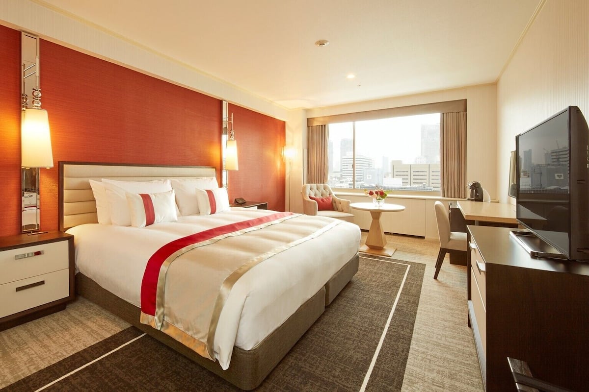 A hotel room with a large bed, a desk, a chair, a TV, and a window with a city view.