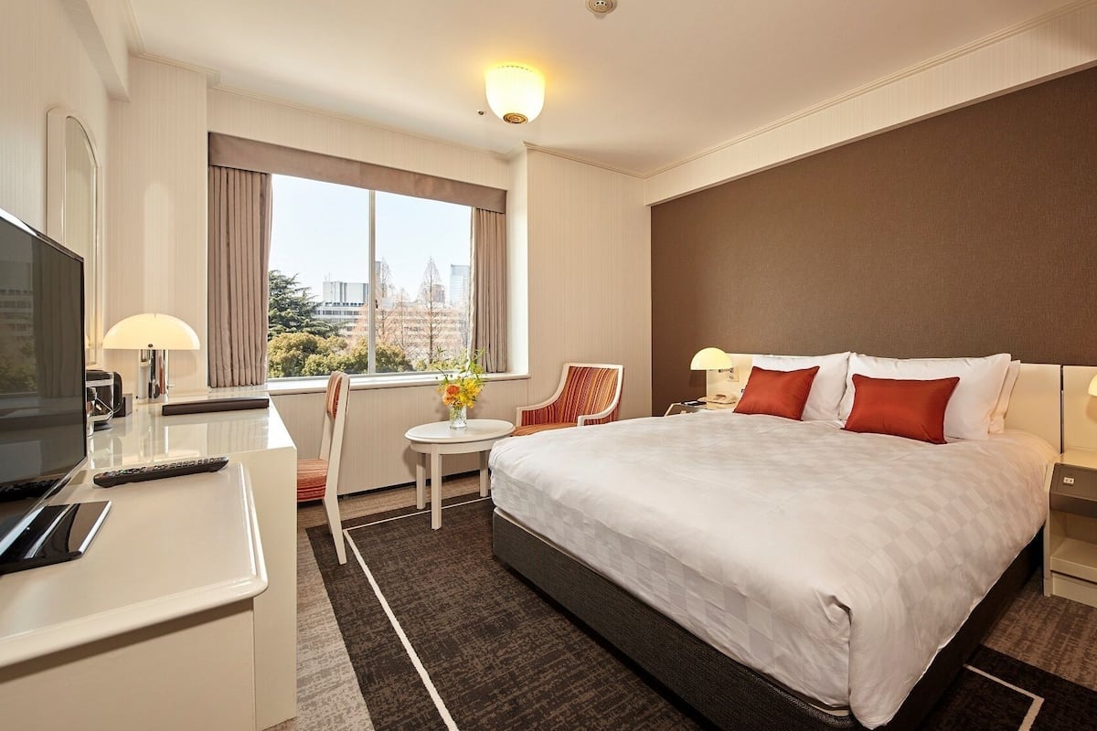 A hotel room with a bed, a desk, a chair, a TV, and a window with a city view.