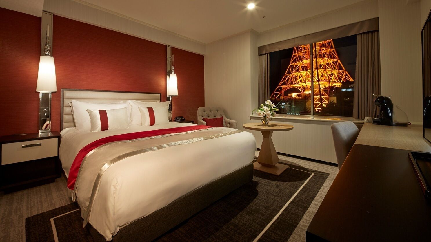A hotel room with a large bed, a desk, a chair, a small table, and a window with a view of a lit structure.