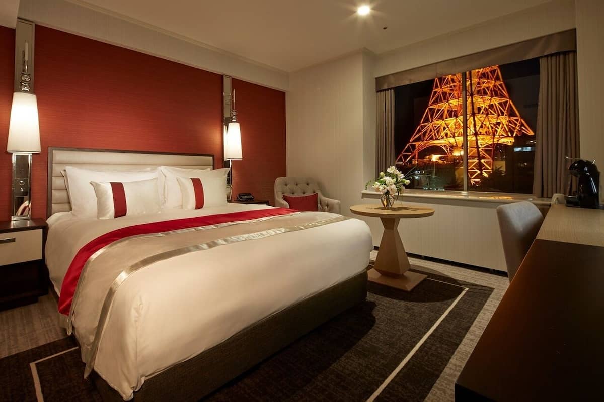 A hotel room with a large bed, a desk, a chair, a small table, and a window with a view of a lit structure.