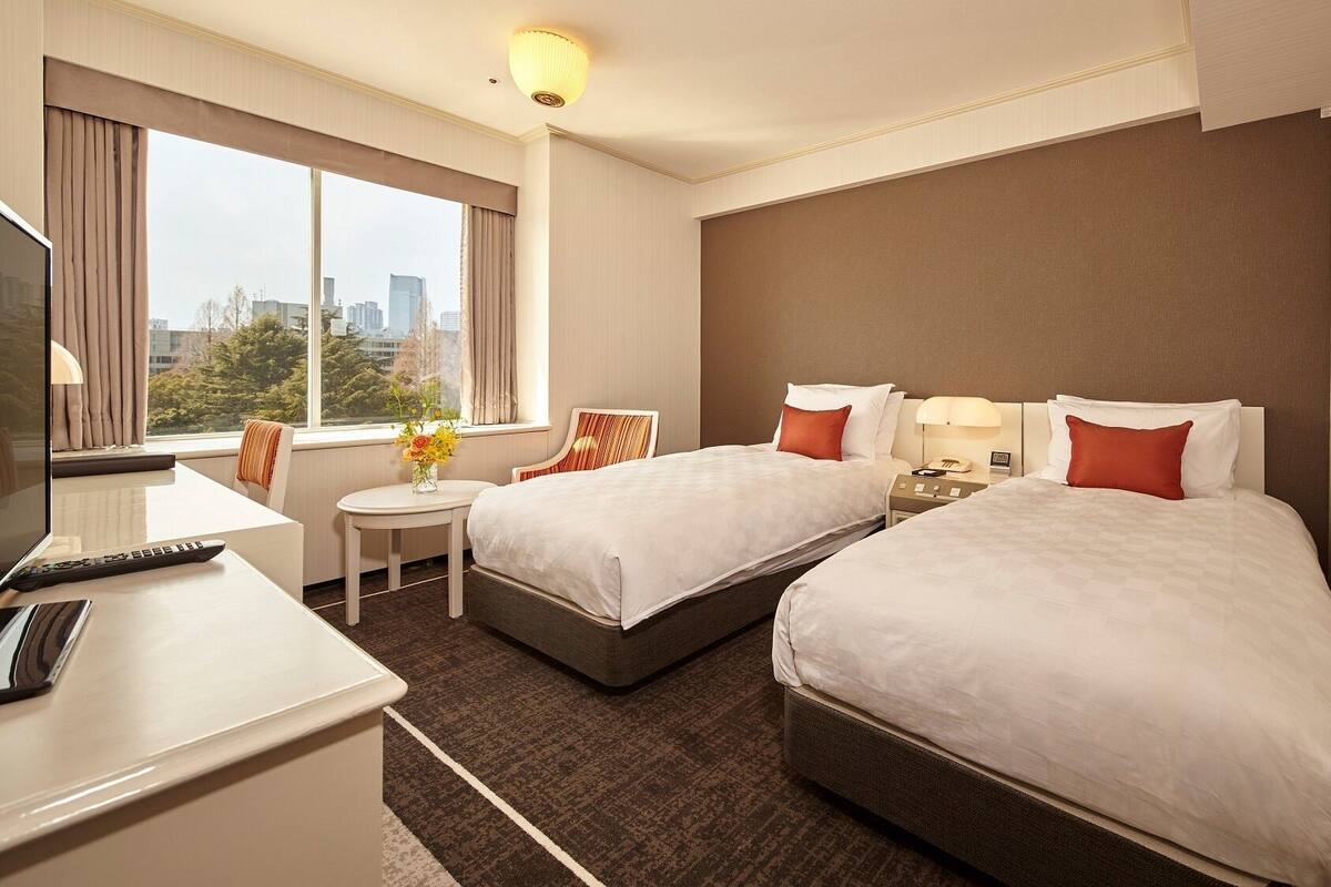 A hotel room with two beds, a desk, a chair, a small table with a vase of flowers, and a window with a city view.