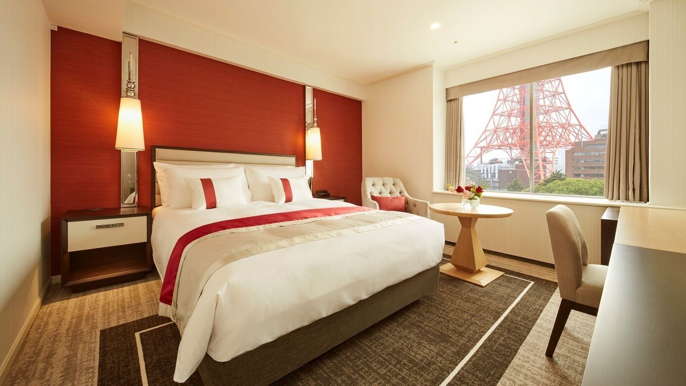A hotel room with a large bed, bedside tables, a round table, a chair, and a window with a view of a red tower.